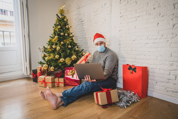 Happy man with face mask home alone in lockdown celebrating virtual christmas video calling family and friend. Virtual holiday gathering online due to coronavirus New normal and social distancing. - 写真・画像
