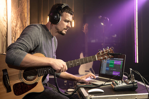 Um guitarrista masculino com fones de ouvido conectados a um mixer de som durante a gravação de som, trabalha com um laptop em um programa de gravação contra um fundo turvo. - Foto, Imagem