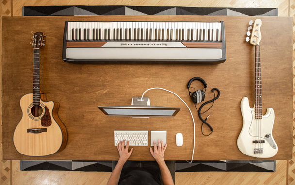 Composition of acoustic guitar, white bass guitar, musical keys, a man at the computer and headphones on a large wooden table. - Foto, Imagen