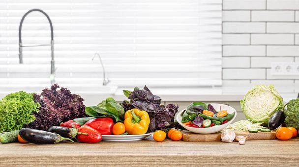 Świeże warzywa w trakcie przygotowywania sałatki na drewnianym stole na tle nowoczesnego wnętrza kuchni. - Zdjęcie, obraz