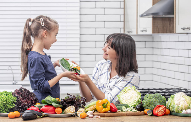 Młoda matka z córką z wieloma warzywami na tle wnętrza nowoczesnej, lekkiej kuchni. - Zdjęcie, obraz