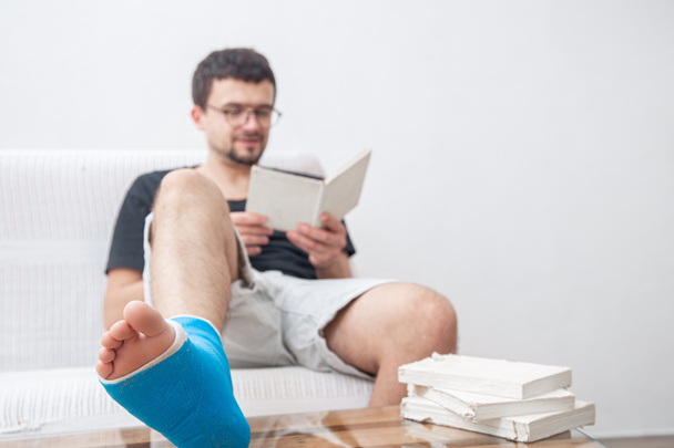 Άνδρας με σπασμένο πόδι σε μπλε νάρθηκα για τη θεραπεία τραυματισμών από διάστρεμμα αστραγάλου διαβάζοντας βιβλία στο σπίτι αποκατάστασης. - Φωτογραφία, εικόνα