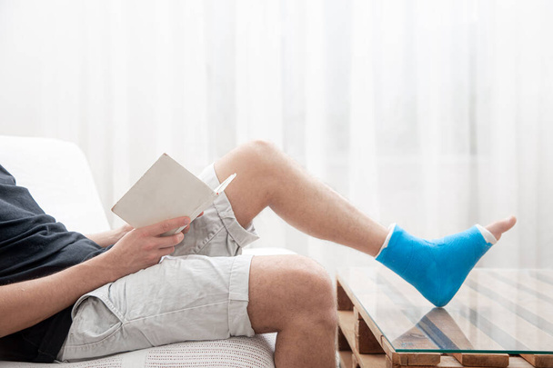 Un hombre con una pierna rota en un yeso lee libros sobre un fondo claro del interior de la habitación. - Foto, Imagen
