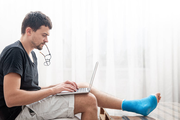 Jovem com uma perna quebrada em tala azul para o tratamento de lesões e entorse de tornozelo está usando um laptop em casa. - Foto, Imagem