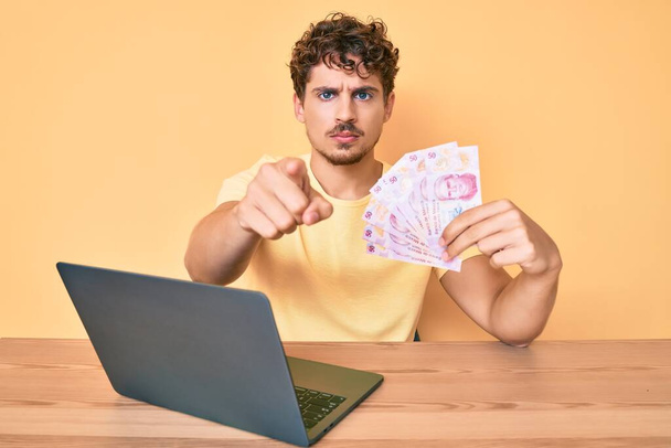 Young caucasian man with curly hair sitting on the table working with laptop and holding mexican pesos banknotes pointing with finger to the camera and to you, confident gesture looking serious  - Photo, image
