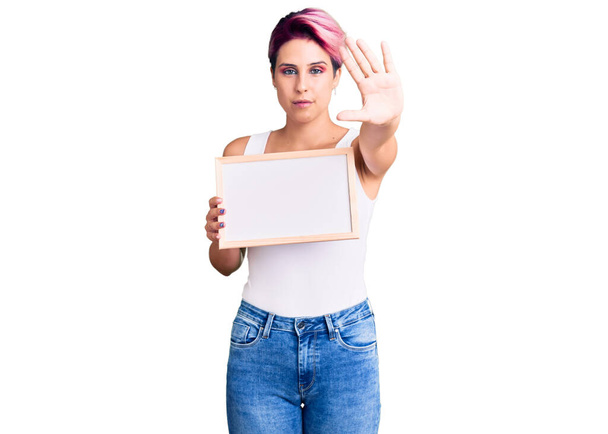 Jonge mooie vrouw met roze haar met lege witte krijtbord met open hand doen stop teken met ernstige en zelfverzekerde uitdrukking, verdediging gebaar  - Foto, afbeelding