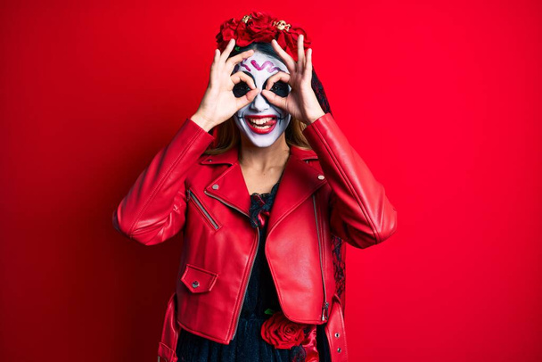 Жінка, одягнена в день мертвого костюма над червоним, робить нормальний жест, як біноклі, що стирчать язиком, очі, дивлячись крізь пальці. божевільний вираз
.  - Фото, зображення