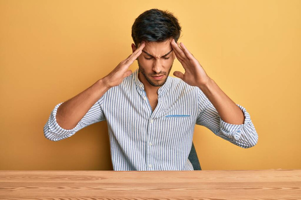 Junger gutaussehender Mann in lässiger Kleidung, der mit der Hand auf dem Kopf am Tisch sitzt, Kopfschmerzen wegen Stress. Migräne.  - Foto, Bild
