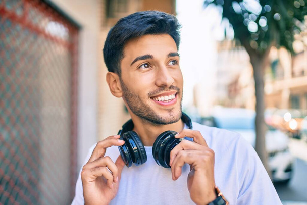 Joven latino sonriendo feliz escuchando música usando auriculares en la ciudad. - Foto, Imagen