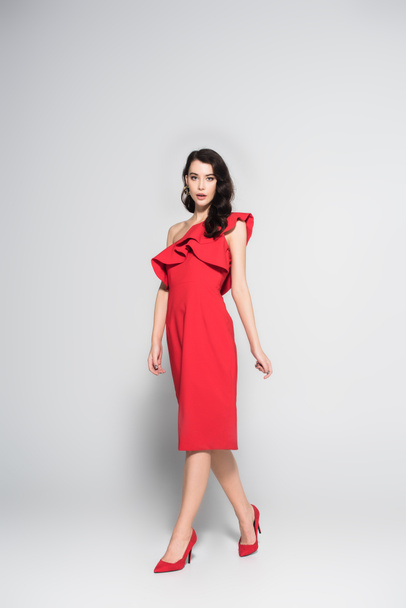 Brunette woman in red dress and heels looking at camera on grey background - Foto, Bild