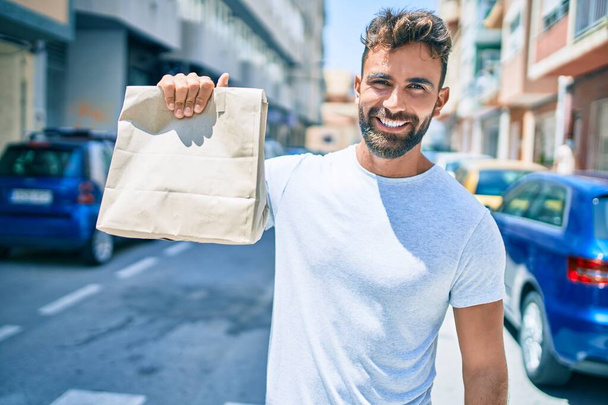 Νεαρός Ισπανός χαμογελάει χαρούμενος κρατώντας μια χάρτινη σακούλα που περπατάει στην πόλη. - Φωτογραφία, εικόνα