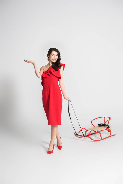Cheerful woman in red dress and heels holding sleigh on grey background - Photo, Image