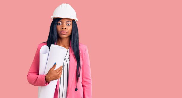 Νεαρή Αφρο-Αμερικανίδα γυναίκα φορώντας καπέλο αρχιτέκτονα κρατώντας σχέδια σκεπτόμενη στάση και νηφάλια έκφραση δείχνοντας αυτοπεποίθηση  - Φωτογραφία, εικόνα