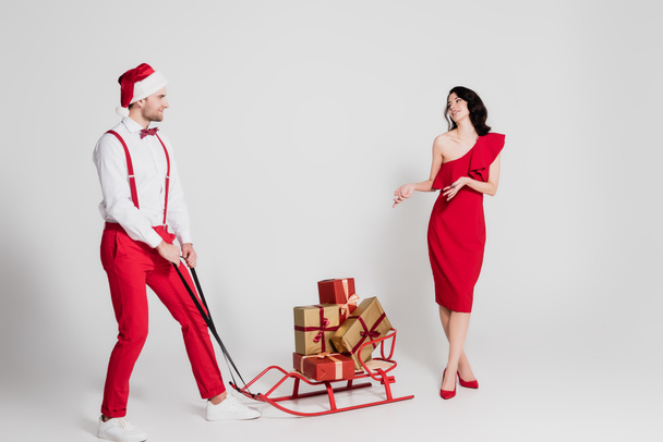 Man in santa hat standing near sleigh with presents and smiling woman in dress on grey background  - Foto, Bild