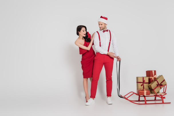 Aufgeregte Frau steht neben lächelndem Mann mit Weihnachtsmütze und deutet auf Schlitten mit Geschenken auf grauem Hintergrund  - Foto, Bild