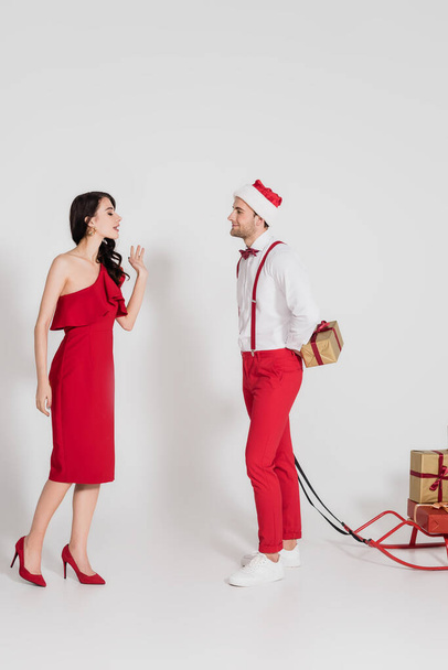 Brunette woman in red dress standing near man in santa hat hiding present and sleigh on grey background  - Foto, Bild