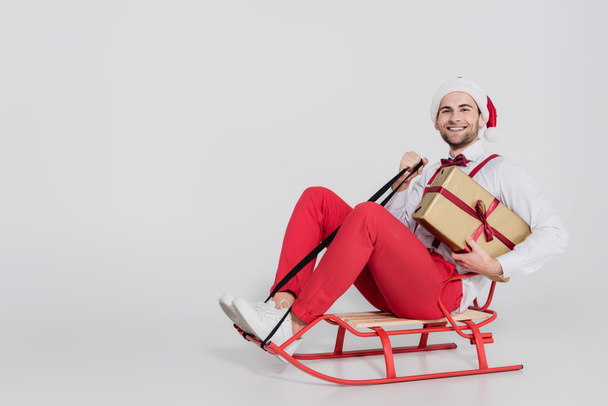 Cheerful man in santa hat holding present while sitting on sleigh on grey background  - Photo, Image