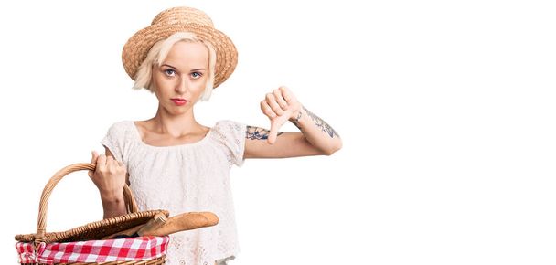 Mujer rubia joven con tatuaje con sombrero de verano y celebración de picnic cesta de mimbre con pan con la cara enojada, signo negativo que muestra aversión con los pulgares hacia abajo, concepto de rechazo  - Foto, Imagen