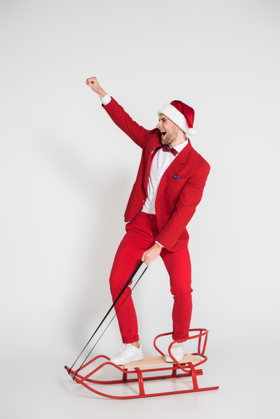 Cheerful man in suit and santa hat standing on sleigh on grey background  - Φωτογραφία, εικόνα