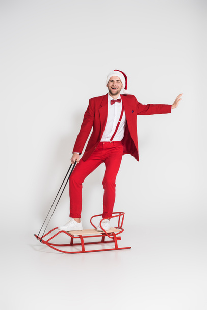 Smiling man in santa hat and red suit looking at camera while standing on sleigh on grey background  - Foto, imagen