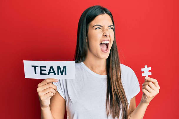 Young brunette woman holding team paper and piece of puzzle as teamwork angry and mad screaming frustrated and furious, shouting with anger. rage and aggressive concept.  - Photo, Image