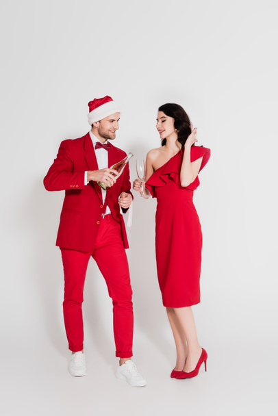 Smiling man in suit and santa hat holding bottle of champagne near woman with glass on grey background  - Foto, imagen