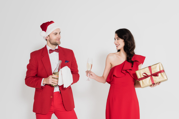 Smiling woman holding glass and gift near man in santa hat with bottle of champagne on grey background  - Foto, imagen