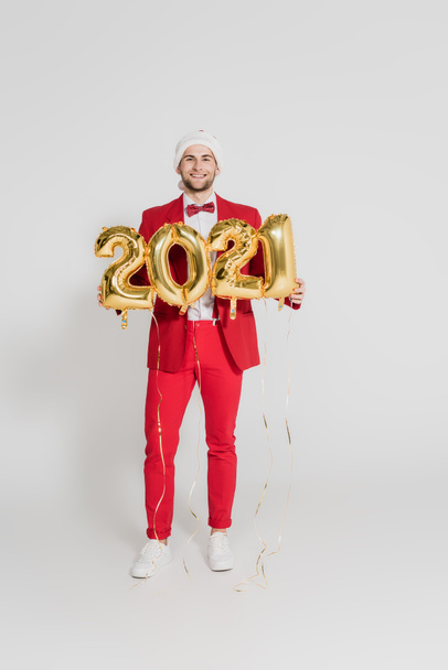 Glimlachende man in santa hoed met ballonnen in de vorm van 2021 nummers op grijze achtergrond  - Foto, afbeelding