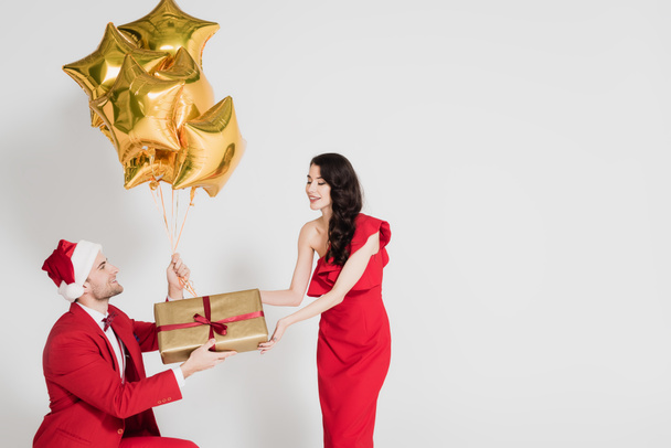 Man in santa hat holding golden balloons and gift near smiling woman on grey background  - Foto, imagen