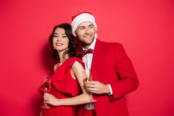 Smiling man in santa hat holding glass of champagne near girlfriend on red background  - Фото, зображення