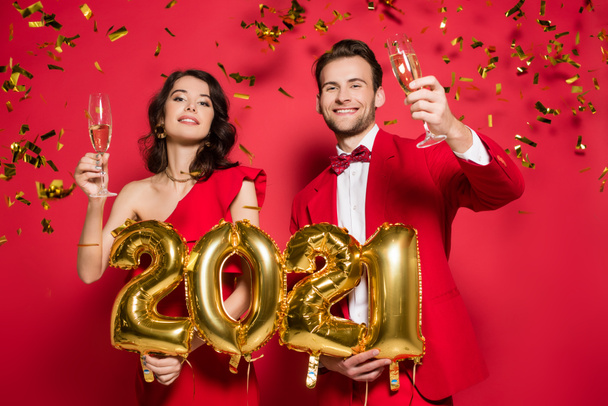 Smiling couple with glasses of champagne holding balloons in shape of 2021 numbers under falling confetti on red background  - Photo, image