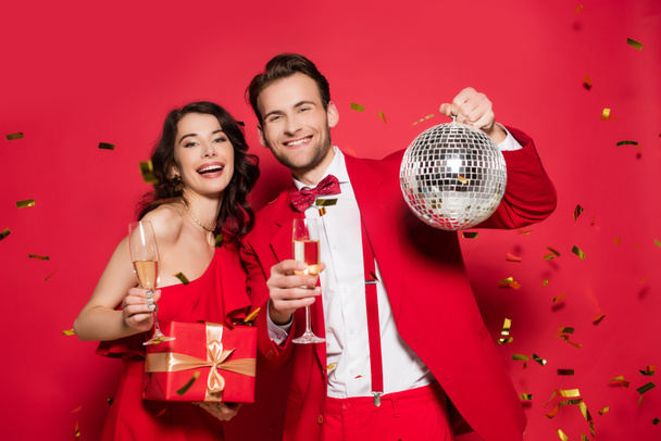 Stilvolles Paar mit Champagnergläsern, Geschenk- und Discokugel unter goldenem Konfetti auf rotem Hintergrund  - Foto, Bild