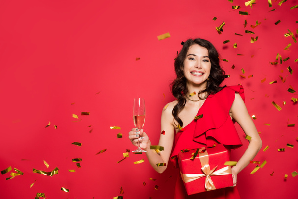 Cheerful woman in dress holding present and glass of champagne under golden confetti on red background  - Foto, imagen