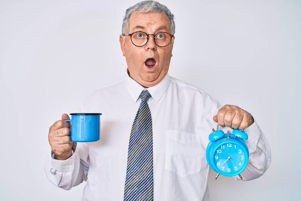 Hombre mayor de pelo gris con ropa de negocios sosteniendo el despertador y la taza de café en la cara de choque, mirando escéptico y sarcástico, sorprendido con la boca abierta  - Foto, imagen