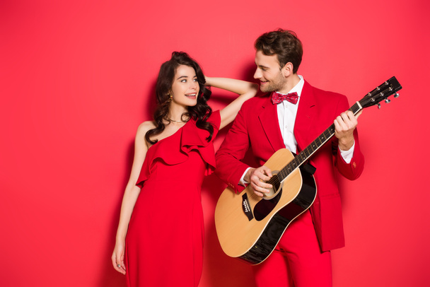 Smiling woman in dress looking at boyfriend playing acoustic guitar on red background  - Fotoğraf, Görsel