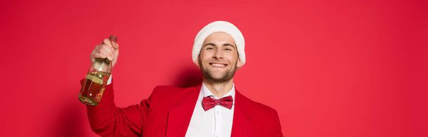 Cheerful man in santa hat holding bottle of champagne on red background, banner  - Foto, Imagen