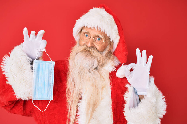 Viejo hombre mayor con disfraz de Santa Claus sosteniendo máscara de seguridad haciendo signo ok con los dedos, sonriendo gesto amistoso excelente símbolo  - Foto, imagen