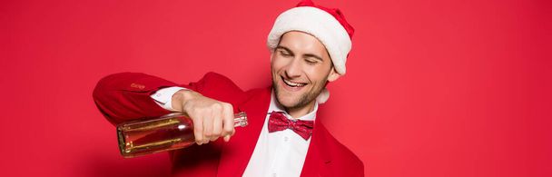 Cheerful man in santa hat holding bottle of champagne on red background, banner  - Photo, image
