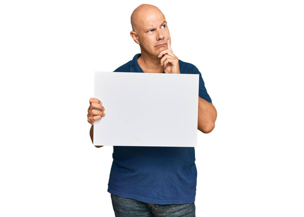 Лысый мужчина средних лет держит пустой баннер серьезное лицо думая о вопросе с рукой на подбородке, вдумчивый о запутанной идеи  - Фото, изображение