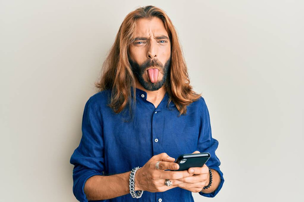 Uzun saçlı ve sakallı çekici bir adam akıllı telefon kullanıyor ve komik bir ifadeyle dilini dışarı çıkarıyor..  - Fotoğraf, Görsel
