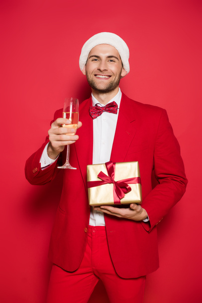 Smiling man in santa hat holding present and glass of champagne on red background  - Valokuva, kuva