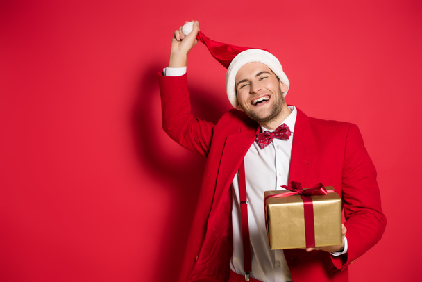 Cheerful man holding santa hat and present on red background  - Фото, зображення