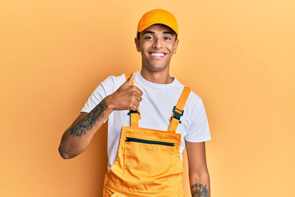 Young handsome african american man wearing handyman uniform over yellow background doing happy thumbs up gesture with hand. approving expression looking at the camera showing success.  - Photo, Image