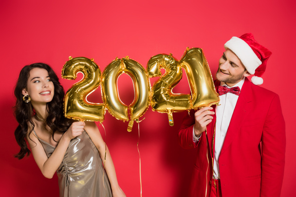 Stylish woman holding balloons in shape of 2021 numbers near boyfriend in santa hat on red background   - Фото, зображення