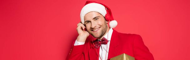Noel Baba şapkalı şık bir adam kırmızı arka planda kulaklıkla müzik dinliyor.  - Fotoğraf, Görsel