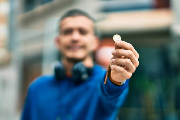 Young hispanic man using headphones holding 1 euro coin at the city - Valokuva, kuva