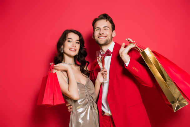 Hombre alegre en traje sosteniendo bolsas de compras cerca de novia con copa de champán sobre fondo rojo - Foto, imagen