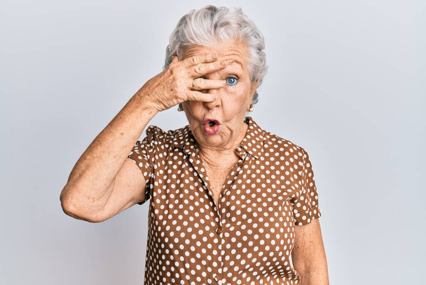 Senior harmaa tukka nainen yllään rento vaatteita kurkistaa shokki peittää kasvot ja silmät kädellä, katselee sormien kautta hämmentynyt ilme.  - Valokuva, kuva