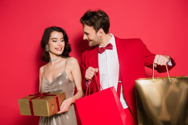 Smiling man holding shopping bags near girlfriend with gift on red background - Foto, Bild