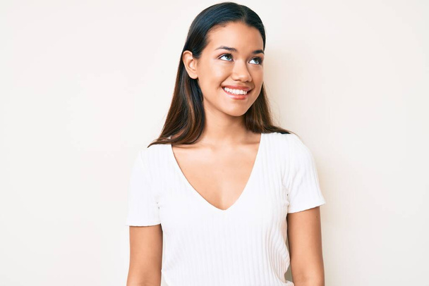 若いです美しいですラテン女の子身に着けていますカジュアル白いtシャツを身に着けています横に見て,リラックスプロファイルポーズとともに自然な顔と自信笑顔.  - 写真・画像